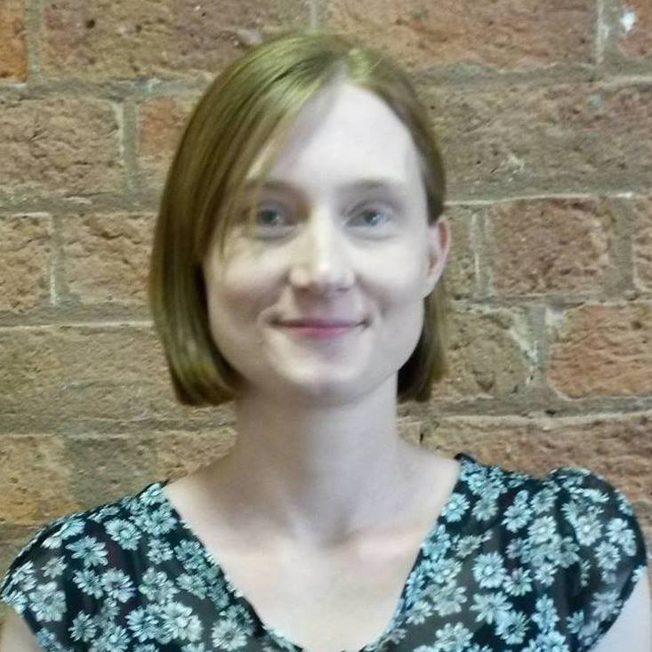 A person with short hair smiles softly while wearing a floral-patterned top, standing against a textured brick wall.