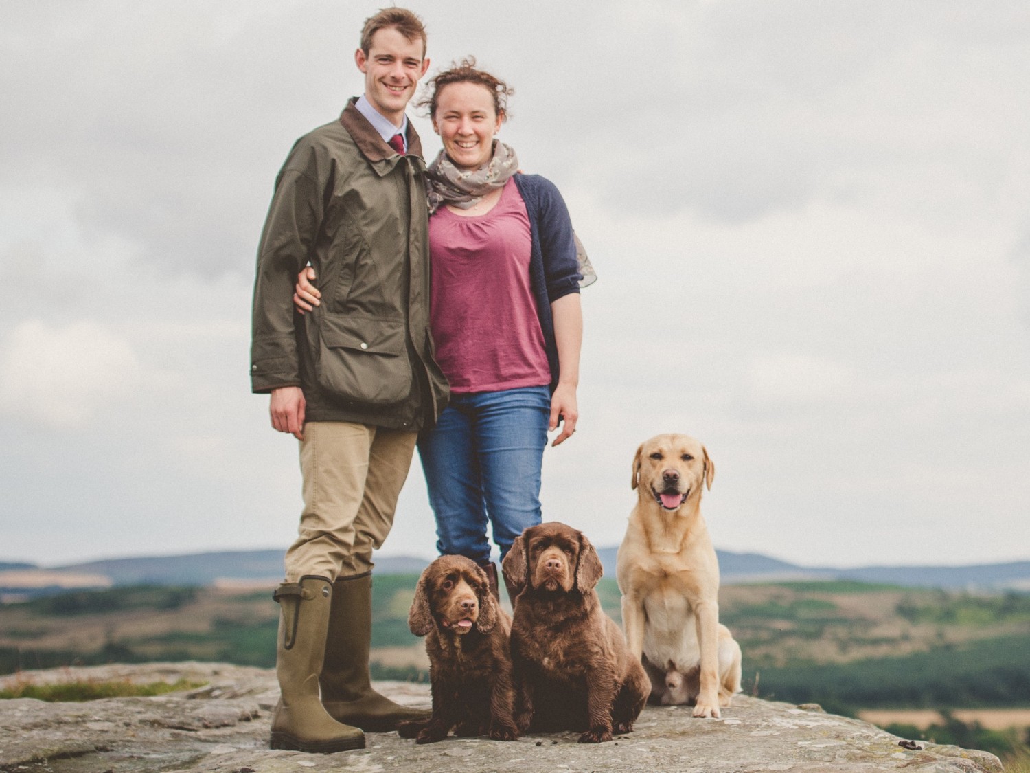 A smiling couple stands arm in arm on a rocky hilltop with three seated dogs. Rolling green hills and a cloudy sky create a serene backdrop.