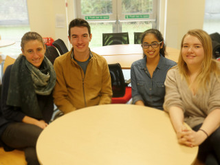Vets-Now-Glasgow-Interns3 Four people smile while seated around a round table in a brightly lit room. A sign on doors behind reads, “Push bar to open.”