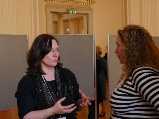Two women are engaged in conversation, standing in a room with tall ceilings and a wooden floor. One woman gestures with her hands, while the other listens attentively.