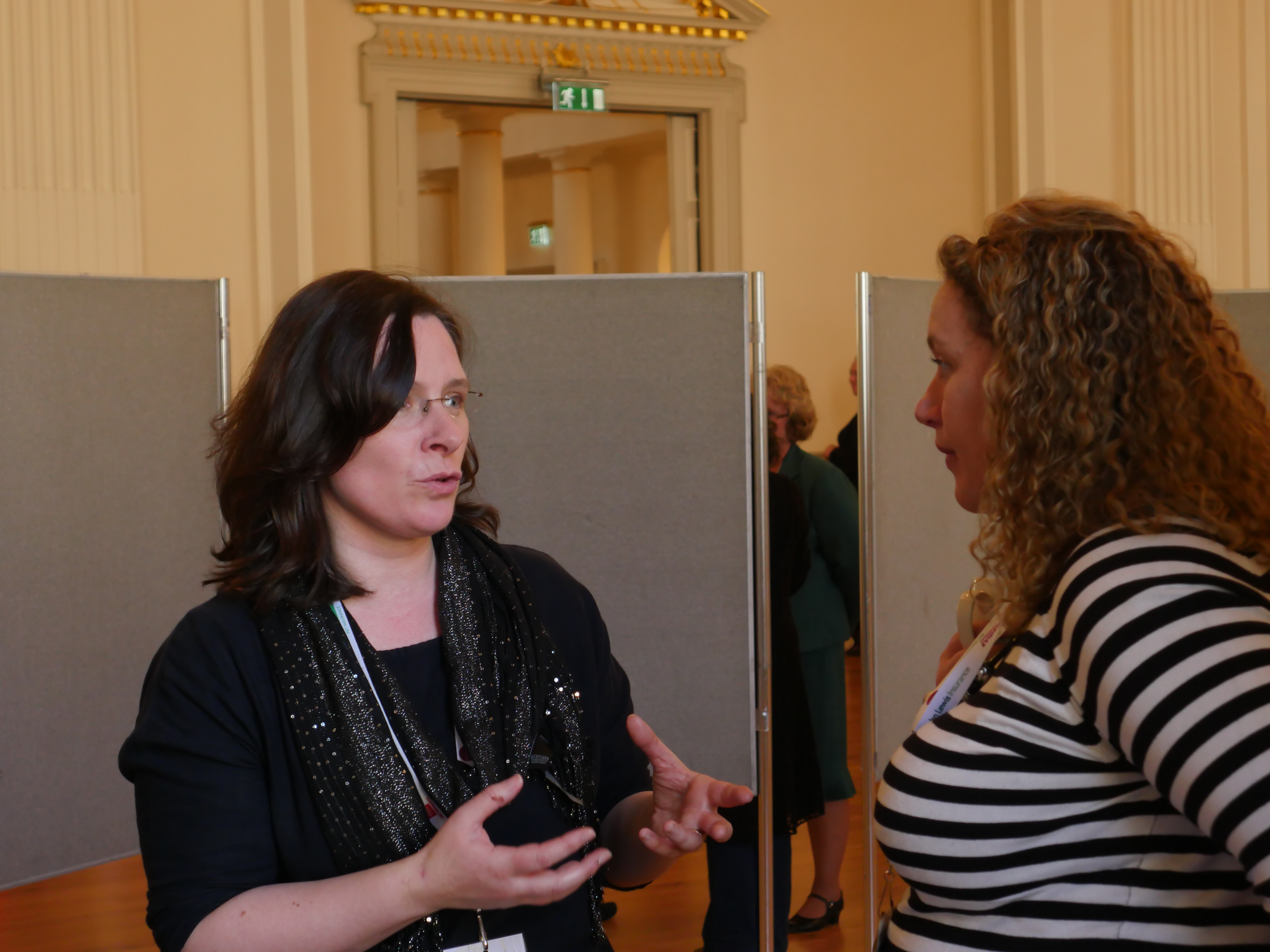 Two women are engaged in conversation, standing in a room with tall ceilings and a wooden floor. One woman gestures with her hands, while the other listens attentively.