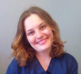 A person smiles warmly, slightly tilting their head, wearing a blue shirt against a plain gray backdrop.