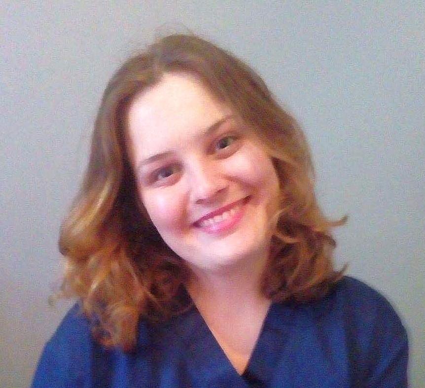 A person smiles warmly, slightly tilting their head, wearing a blue shirt against a plain gray backdrop.