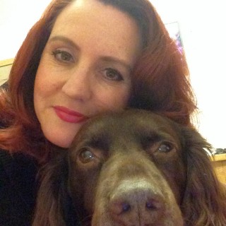 A woman with red hair gently embraces a brown dog, both facing the camera closely in an indoor setting with a soft-lit background.
