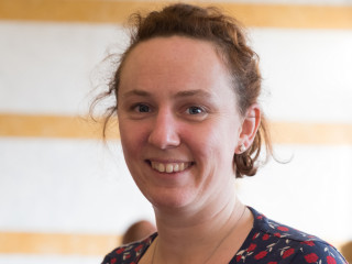 Sara Jackson A person smiling indoors, with light brown hair and floral-patterned clothing. Soft lighting highlights their face, with a blurred, cream and yellow-striped background. No text present.