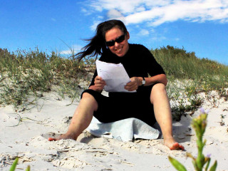Tanja Klein revising in Australia A person wearing sunglasses reads papers while sitting on a sandy beach, surrounded by grasses under a bright blue sky with clouds.