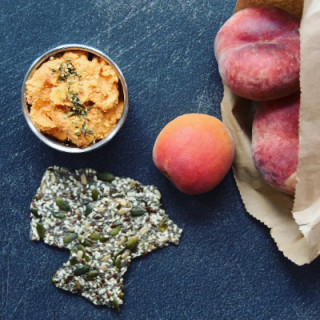 Red pepper hummus A bowl of orange dip garnished with herbs sits beside a seed cracker and a fresh apricot, with peaches partially visible in a brown paper bag, on a dark surface.