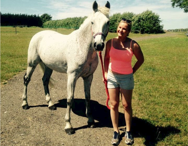 Victoria Mansfield A white horse stands calmly beside a person holding its lead, in a sunny field with a dirt path, surrounded by green grass and trees.