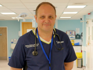 A veterinary surgeon stands smiling in a clinical setting, wearing a stethoscope and a navy uniform with text: 