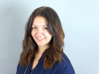 Gill Izzy, senior people services business partner A person with long, wavy brown hair smiles in front of a plain, light gray background, wearing a dark blue top.