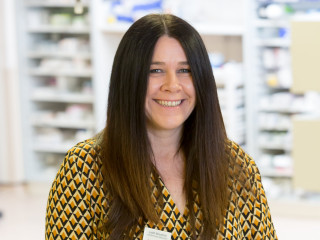 Judith McLetchie Portrait-min A smiling woman with long, straight hair wears a geometric-patterned top and name badge, standing in a room with blurred shelves and various items in the background.