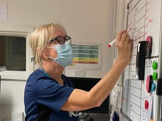 Valerie Munro ACA 3 (1) A healthcare worker wearing a mask and glasses writes on a whiteboard with a red marker in a medical office setting.
