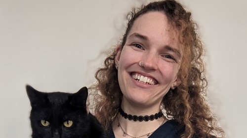 A person with curly hair smiles while holding a calm black cat; they both appear indoors against a plain background.