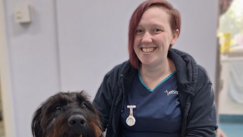 A veterinarian from Vets Now smiling and sitting beside a large black dog, both looking at the camera in a clinic setting.