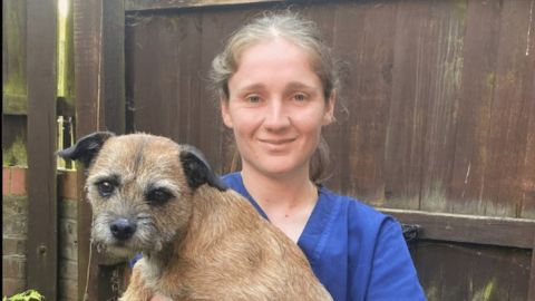 A veterinarian in a Vets Now uniform holding a small brown terrier, standing outdoors by a wooden fence, both looking at the camera.