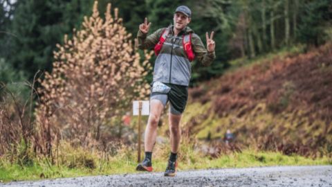 A man running on a gravel trail in an outdoor setting, giving peace signs with both hands, wearing a red backpack and athletic gear