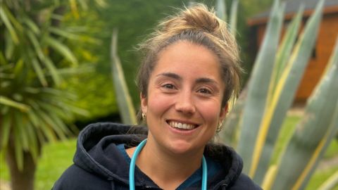 A veterinarian in a Vets Now uniform smiling outdoors, with large plants in the background, conveying a warm and friendly presence.