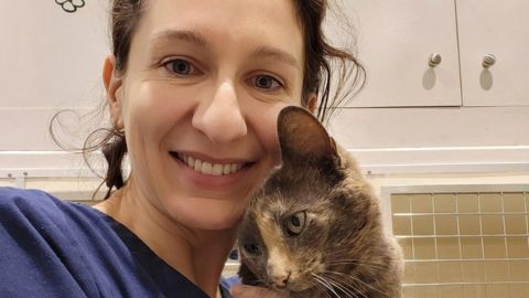 Vets-Now Vet Victoria Lenthall smiling while holding a tortoiseshell cat at a Vets Now clinic. The background shows veterinary cabinets and a cat enclosure.