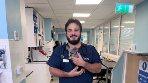 Edward Selvaig in a veterinary clinic setting, holding a small dog and smiling. The clinic background includes medical equipment and supplies.