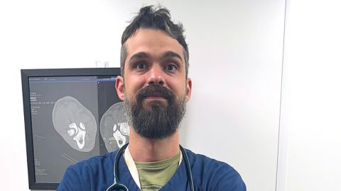 Cristian Mihaita, a veterinarian wearing Vets-Now scrubs and a stethoscope, standing with arms crossed in front of an X-ray display in a clinical setting.