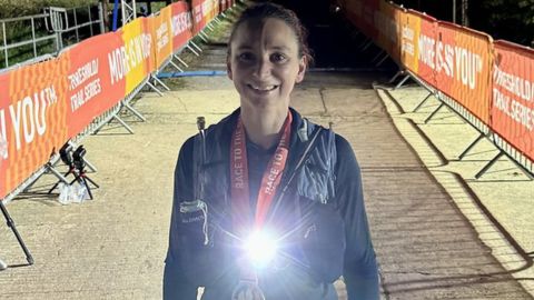 Zara Kennedy smiling at the finish line of an evening trail race, wearing a head torch and finisher’s medal.