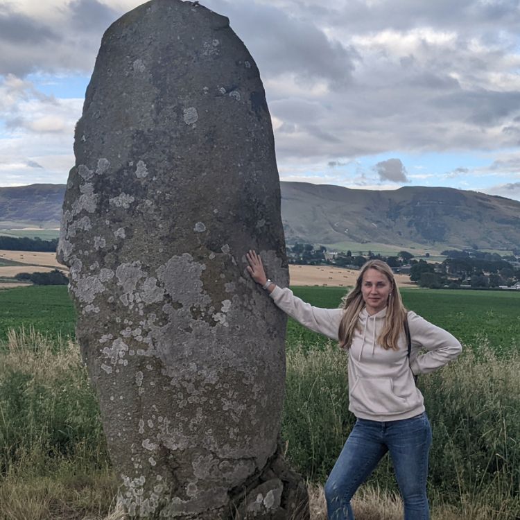 weronika-misna-outdoors-standing-stone Weronika Misna standing beside a large standing stone in a rural field, wearing a hoodie and jeans, with scenic hills in the background.