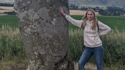 Weronika Misna standing beside a large standing stone in a rural field, wearing a hoodie and jeans, with scenic hills in the background.